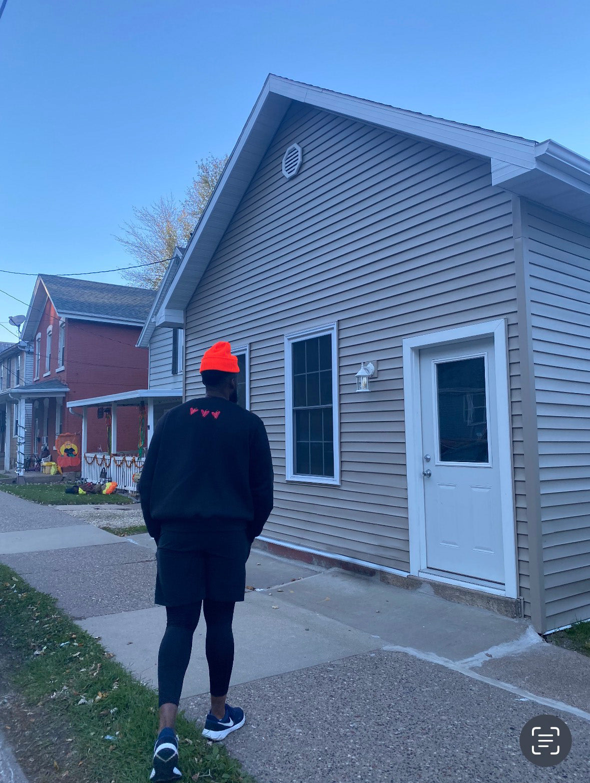 Person wearing a black jacket and orange beanie walking on a sidewalk in front of a house.