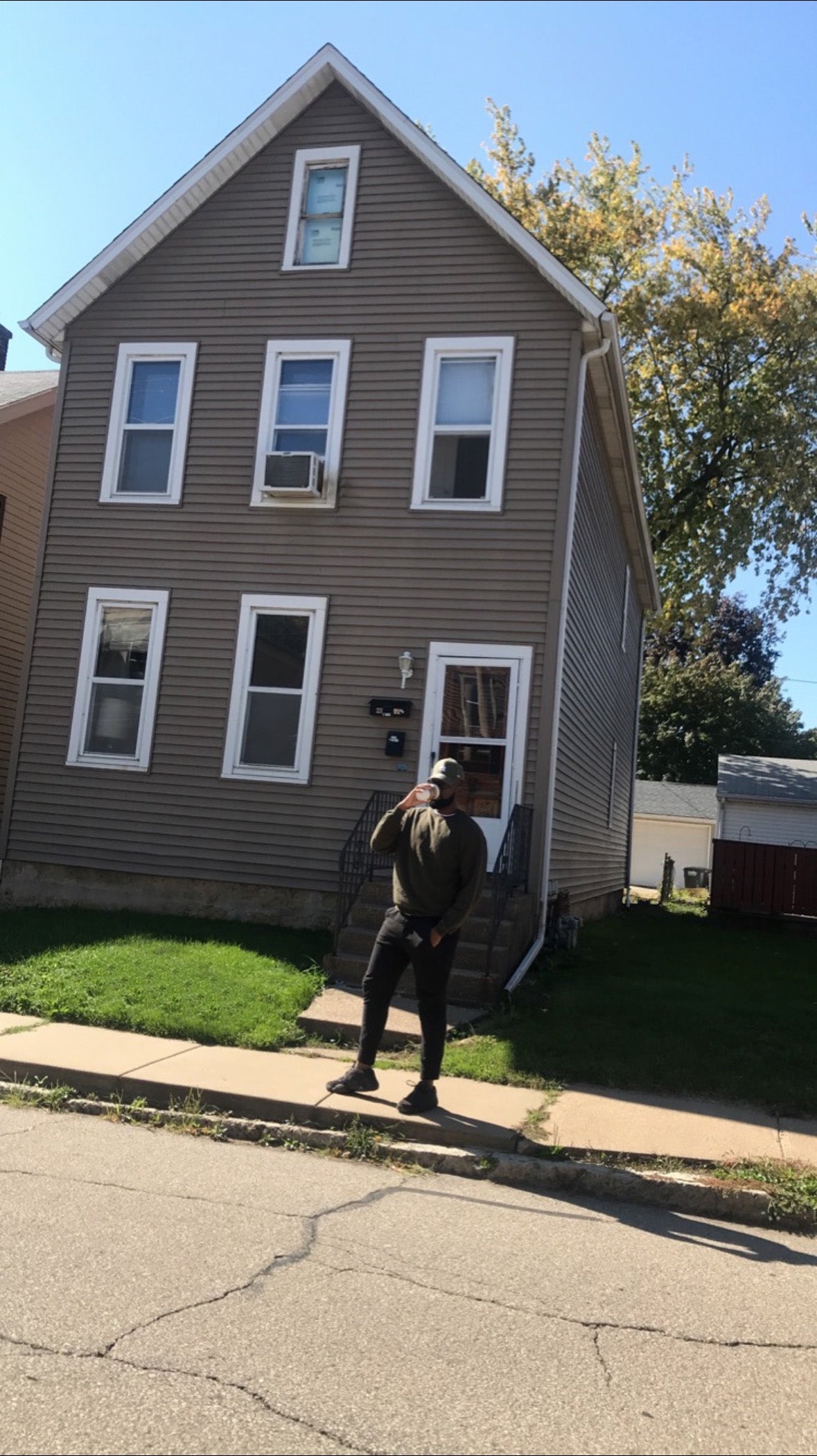 Person standing on a sidewalk in front of a two-story house with a clear blue sky.